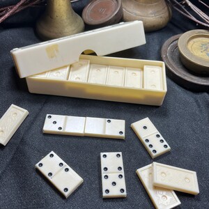 Può includere: Un set di tessere del domino color bianco sporco con punti neri e retro testurizzato, completo di scatola rettangolare coordinata con coperchio scorrevole. Diverse tessere sono disposte all'esterno della scatola, mostrando vari motivi a punti.