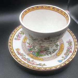 May include: A white teacup and saucer with a floral pattern in shades of yellow, pink, blue, and green. The pattern features a stylized tree with birds and flowers.