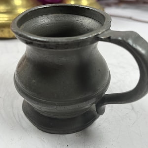 Puede incluir: Una peque&ntilde;a jarra antigua de peltre con asa. La jarra tiene un cuerpo redondeado y una base acampanada. El asa es curva y est&aacute; unida al lateral de la jarra. La jarra es de color gris oscuro y parece antigua.