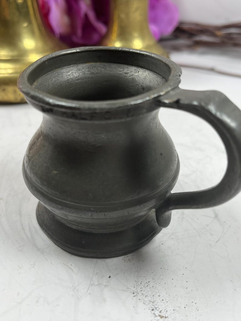 Puede incluir: Una peque&ntilde;a jarra antigua de peltre con asa. La jarra tiene un cuerpo redondeado y una base acampanada. El asa es curva y est&aacute; unida al lateral de la jarra. La jarra es de color gris oscuro y parece antigua.