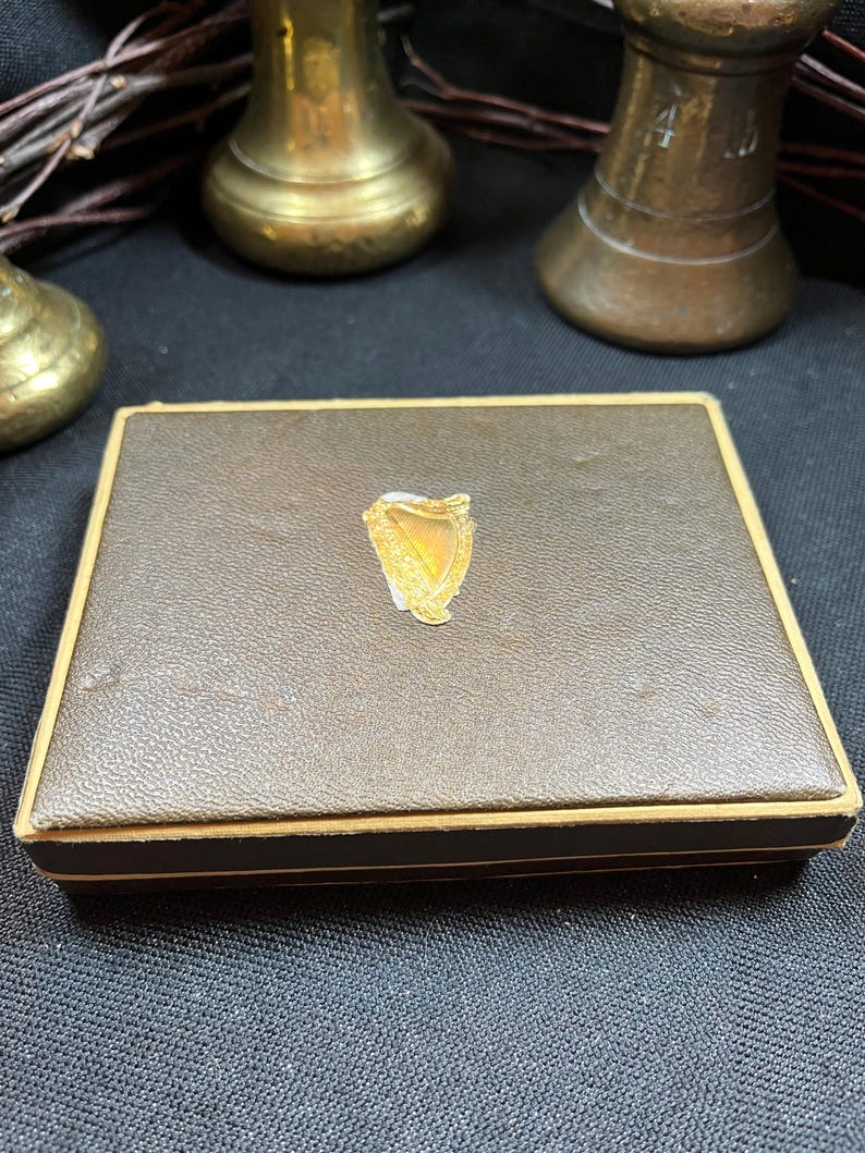 May include: A brown rectangular box with a gold trim and a gold emblem in the center. The box has a textured surface and appears to be made of leather or a similar material. The emblem is a gold harp design.
