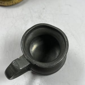 Puede incluir: Una peque&ntilde;a jarra de peltre antigua con asa. La jarra tiene un color gris met&aacute;lico oscuro y un dise&ntilde;o simple y cl&aacute;sico. El interior de la jarra es visible, mostrando un fondo liso y redondeado. El asa est&aacute; unida al lateral de la jarra.