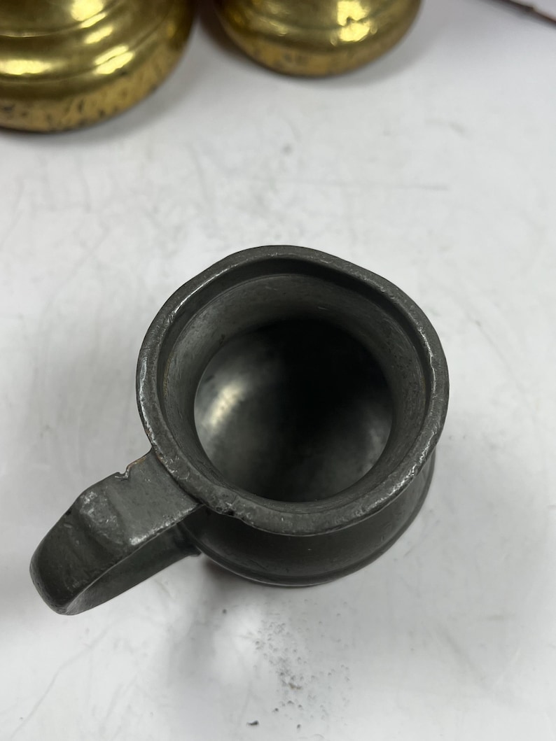 Puede incluir: Una peque&ntilde;a jarra de peltre antigua con asa. La jarra tiene un color gris met&aacute;lico oscuro y un dise&ntilde;o simple y cl&aacute;sico. El interior de la jarra es visible, mostrando un fondo liso y redondeado. El asa est&aacute; unida al lateral de la jarra.