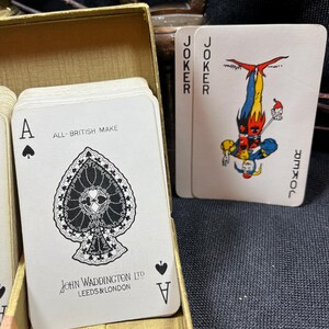 May include: A deck of playing cards in a gold box. The Ace of Spades card is visible, with a detailed black and white design. The text "ALL-BRITISH MAKE" and "JOHN WADDINGTON LTD LEEDS & LONDON" are printed on the card. Two Joker cards are also visible.