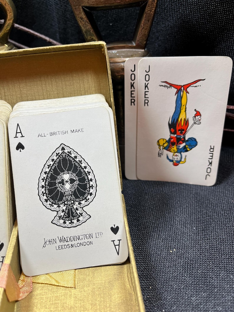 May include: A deck of playing cards in a gold box. The Ace of Spades card is visible, with a detailed black and white design. The text "ALL-BRITISH MAKE" and "JOHN WADDINGTON LTD LEEDS & LONDON" are printed on the card. Two Joker cards are also visible.