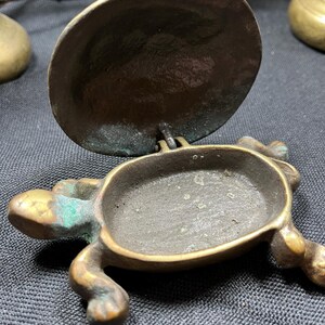 May include: A small, antique brass turtle-shaped container with a hinged lid. The container has a dark, aged patina with hints of green oxidation. The turtle's legs and head are detailed, and the lid is open, revealing the interior.