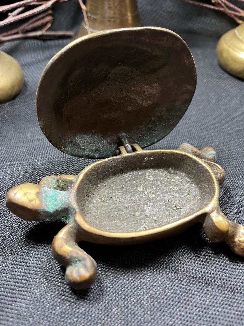 May include: A small, antique brass turtle-shaped container with a hinged lid. The container has a dark, aged patina with hints of green oxidation. The turtle's legs and head are detailed, and the lid is open, revealing the interior.
