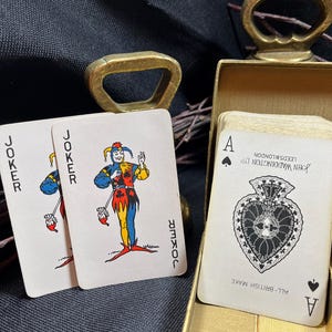 May include: Two Joker playing cards and an Ace of Spades card are displayed, with a gold-colored bottle opener and a gold box in the background. The Joker cards feature a colorful jester design. The Ace of Spades card is from John Waddington Ltd.