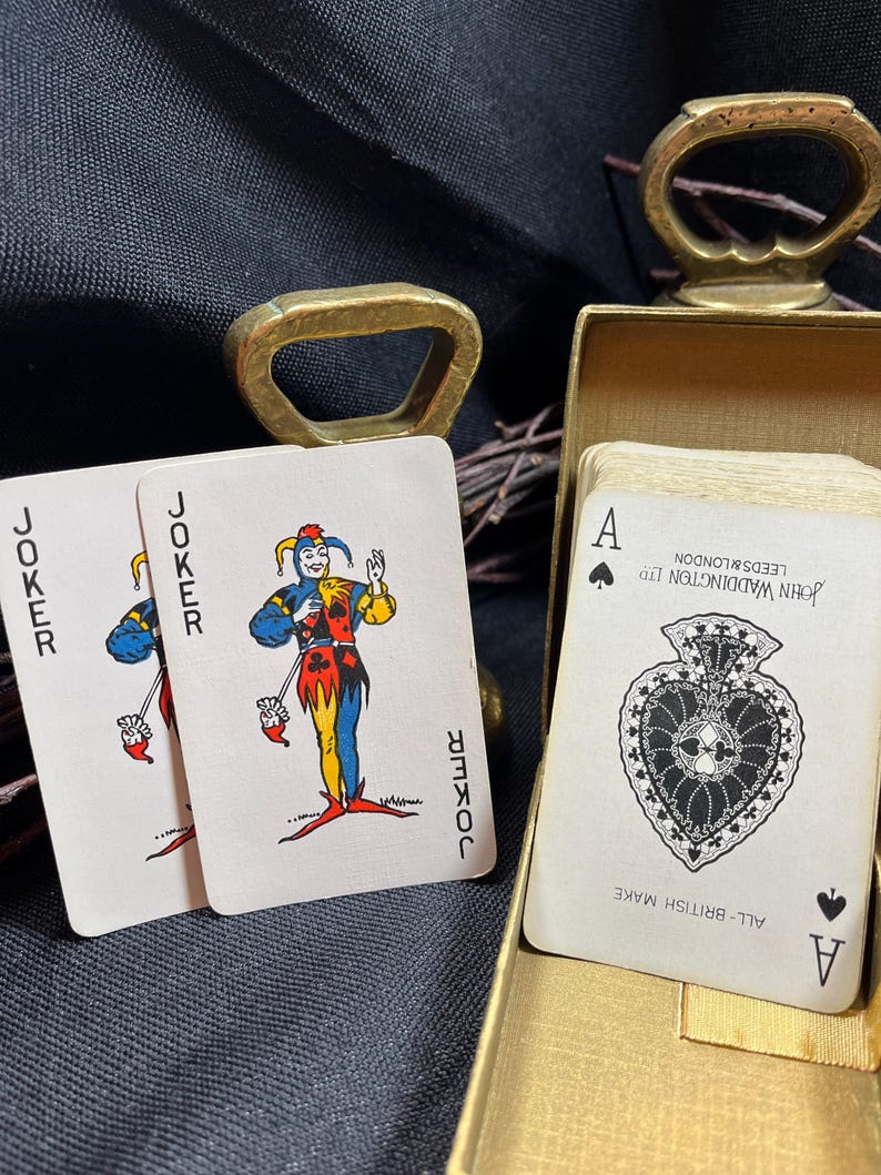 May include: Two Joker playing cards and an Ace of Spades card are displayed, with a gold-colored bottle opener and a gold box in the background. The Joker cards feature a colorful jester design. The Ace of Spades card is from John Waddington Ltd.