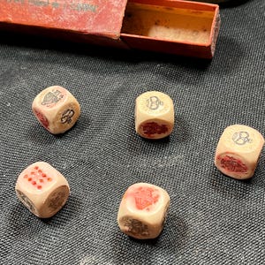 May include: Five vintage ivory-colored dice with red markings and symbols, scattered on a dark surface. A red rectangular box is partially visible in the background. The dice show various symbols and numbers, suggesting a game or collectible item.