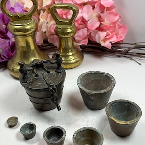 Op de afbeelding: Een verzameling antieke messing en bronzen gewichten en meetinstrumenten. De set bevat twee grote messing gewichten met handvatten, een kleine bronzen weegschaal en verschillende kleine bronzen maatbekers. De items zijn gerangschikt op een wit oppervlak met roze bloemen op de achtergrond.