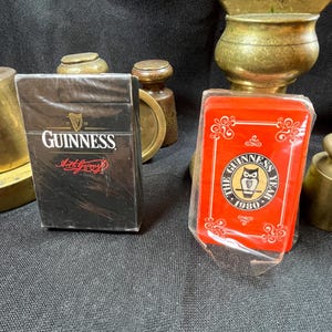 Op de afbeelding: Een zwart pakje Guinness speelkaarten en een rood kaartspel van Guinness in een plastic verpakking. Het zwarte pakje heeft het Guinness-logo en de woorden "Arthur Guinness" in het rood. De rode set heeft een gouden uil-logo en de woorden "The Guinness Year 1980".