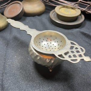 Peut inclure: Une passoire à thé en métal argenté avec un bol perforé et une poignée décorative ornée d'un motif en treillis. La passoire repose sur un petit récipient en métal arrondi et cannelé, doté d'une finition métallique et de petits pieds.