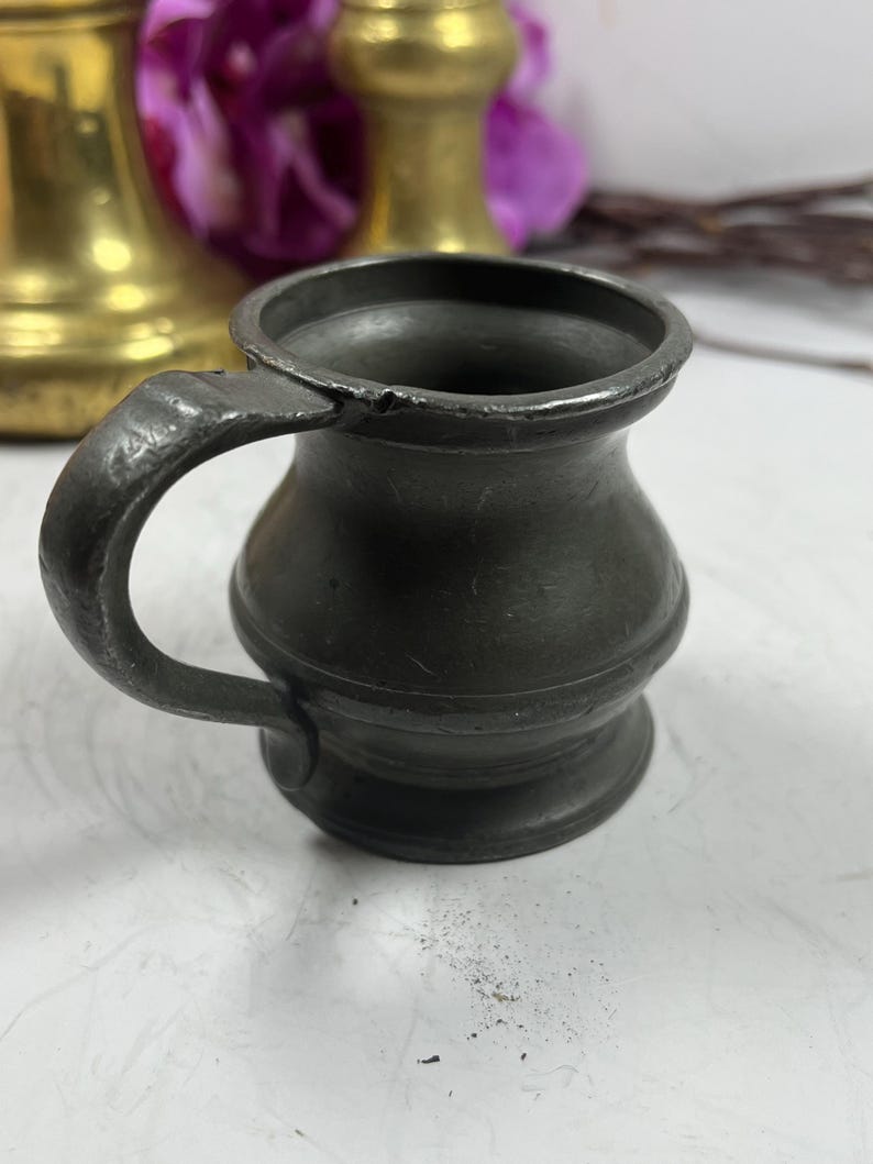 Puede incluir: Una peque&ntilde;a jarra de peltre antigua con un cuerpo redondeado y un asa robusta. La jarra tiene un aspecto oscuro y envejecido, con desgaste visible, lo que sugiere su antig&uuml;edad. El fondo incluye otros objetos de lat&oacute;n y flores moradas.