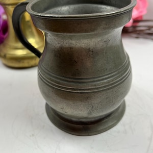 Könnte beinhalten: Ein kleiner, antiker Zinnhumpen mit dunklem Griff. Der Humpen hat einen abgerundeten Körper mit horizontalen Bändern und einem ausgestellten Rand. Der Hintergrund umfasst ein Messingobjekt und rosa Blumen.