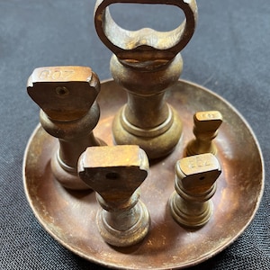 May include: A set of antique brass weights with wooden tops, displayed on a circular metal tray. The weights are of different sizes and are marked with numbers. The tray and weights show signs of age, indicating they are vintage.