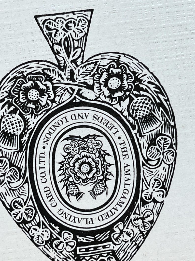May include: Black and white playing card illustration featuring a heart shape with floral and thistle designs. The text reads "THE AMALGAMATED PLAYING CARD CO. LTD. LONDON AND LEEDS". The spade symbol is at the top.