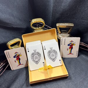 May include: A deck of playing cards with the Ace of Spades and Joker cards visible, presented in a gold box tied with a yellow ribbon. The image also shows three gold-colored bottle openers and a ring.