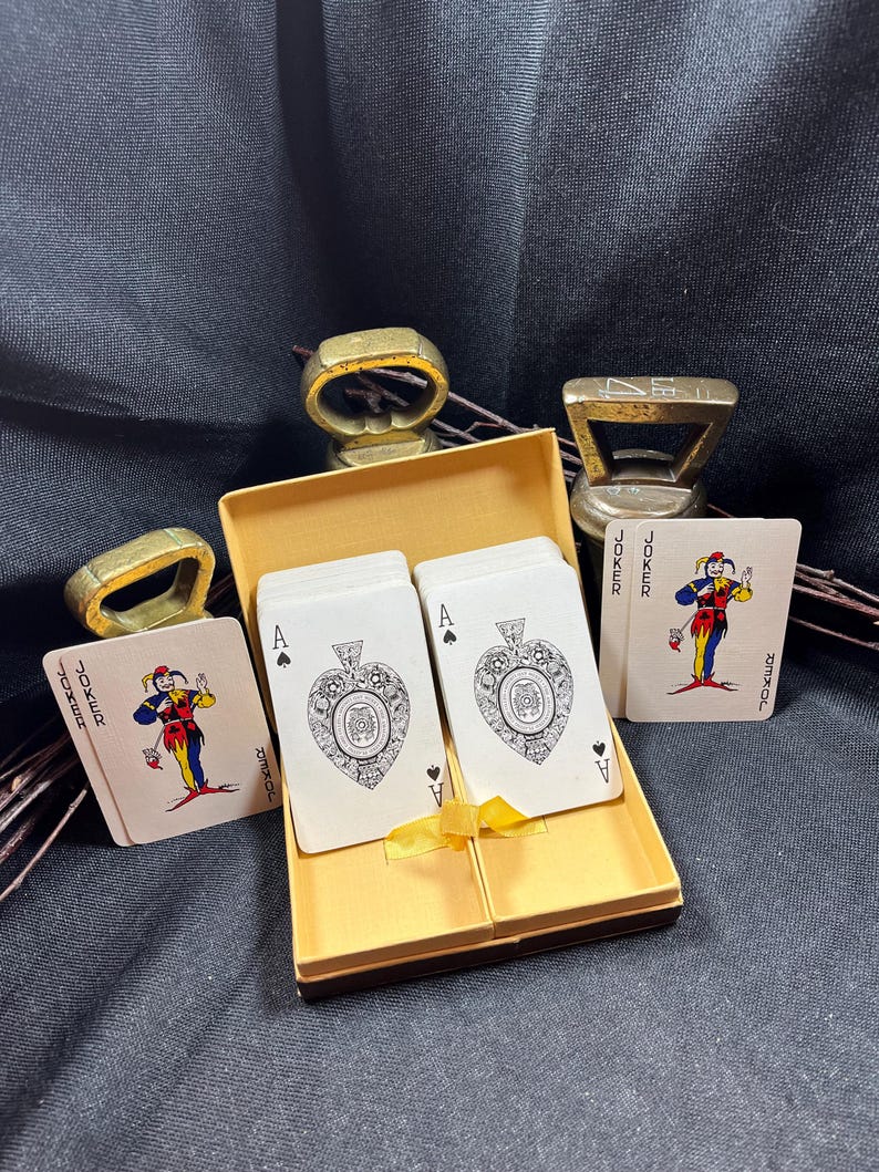 May include: A deck of playing cards with the Ace of Spades and Joker cards visible, presented in a gold box tied with a yellow ribbon. The image also shows three gold-colored bottle openers and a ring.