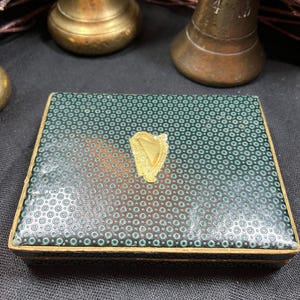 May include: A rectangular box with a teal and black dotted pattern on the lid. A gold-colored harp emblem is centered on the lid. The box has gold-colored trim and is set against a dark background.