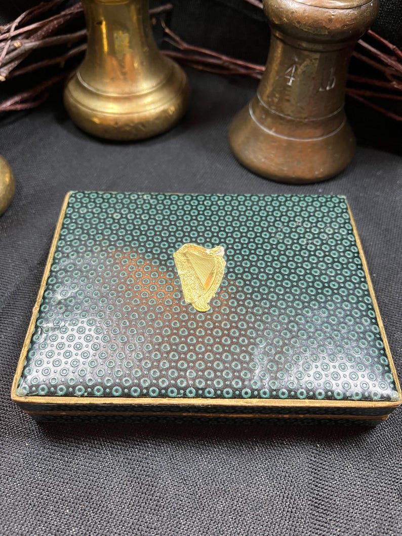 May include: A rectangular box with a teal and black dotted pattern on the lid. A gold-colored harp emblem is centered on the lid. The box has gold-colored trim and is set against a dark background.