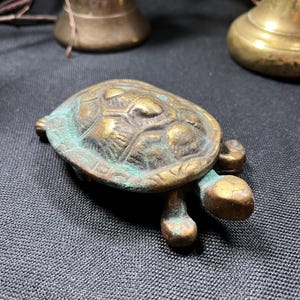May include: A small, antique brass turtle figurine with a weathered, verdigris finish. The turtle has a detailed shell and stubby legs. The figurine is on a dark gray textured surface, with brass bells in the background.