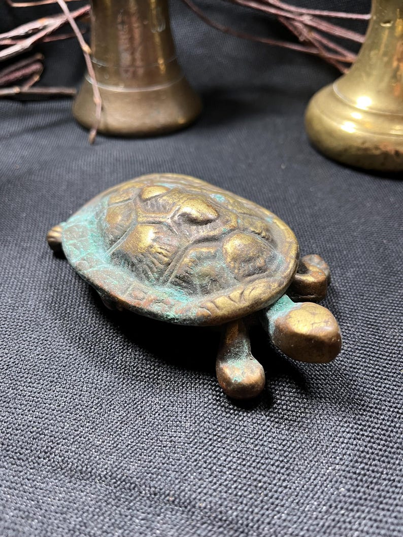 May include: A small, antique brass turtle figurine with a weathered, verdigris finish. The turtle has a detailed shell and stubby legs. The figurine is on a dark gray textured surface, with brass bells in the background.