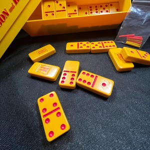 Könnte beinhalten: Ein gelbes Vintage-Dominoset mit roten Punkten, verstreut auf einer dunklen Oberfläche. Das Set enthält eine gelbe Schale und eine Box mit rotem Text. Einige Dominosteine sind in einer Reihe angeordnet, andere verstreut.