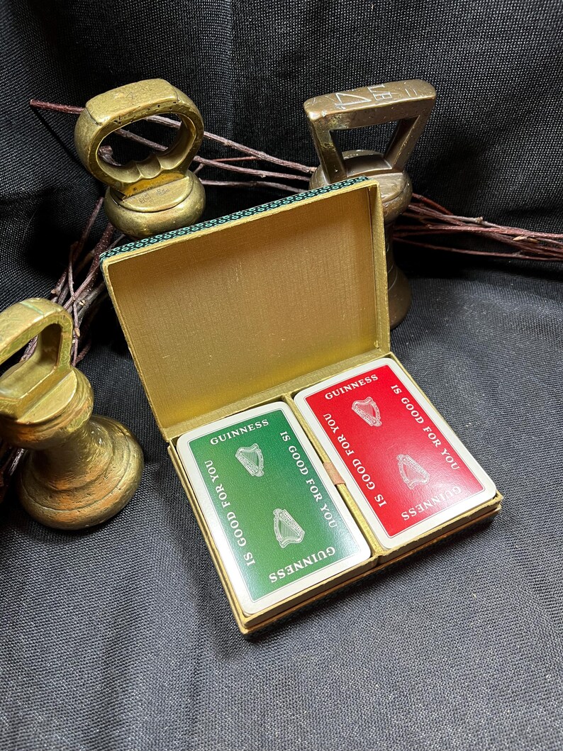 May include: A vintage gold-colored playing card box with two decks of cards. Each card has the Guinness logo and the text "IS GOOD FOR YOU". The box is open, revealing the green and red card backs.