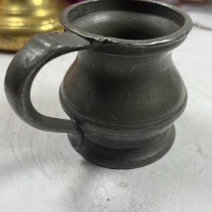 Puede incluir: Una peque&ntilde;a jarra de peltre antigua con asa. La jarra tiene un cuerpo redondeado y un borde acampanado. El asa es curva y est&aacute; unida al lateral. El peltre tiene un aspecto oscuro y envejecido, lo que sugiere su antig&uuml;edad.
