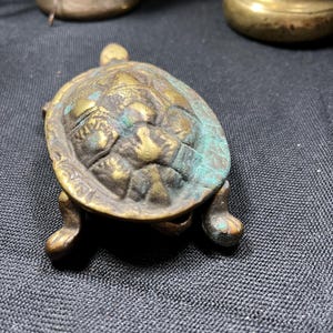 May include: A small, antique brass turtle figurine with a textured shell and four stubby legs. The turtle has a weathered appearance with hints of green patina. The figurine is a decorative item.