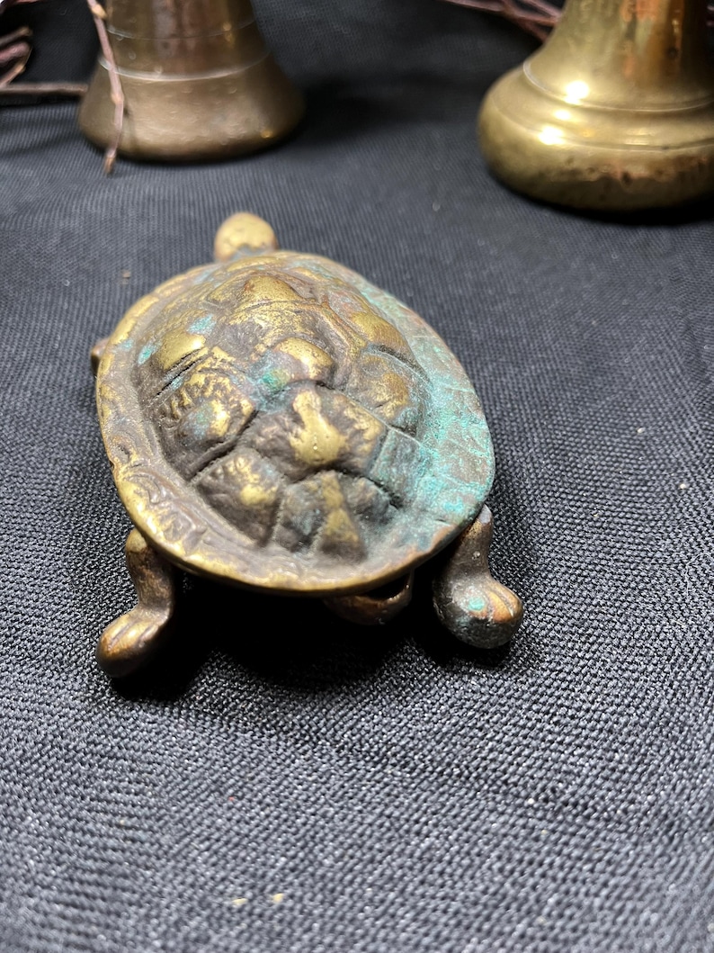 May include: A small, antique brass turtle figurine with a textured shell and four stubby legs. The turtle has a weathered appearance with hints of green patina. The figurine is a decorative item.