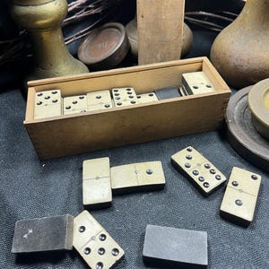 Peut inclure: Un jeu de dominos rectangulaires avec des faces couleur crème et des points noirs, rangé dans une boîte en bois rectangulaire. Plusieurs dominos sont disposés hors de la boîte, montrant les faces à points et les dos noirs unis.