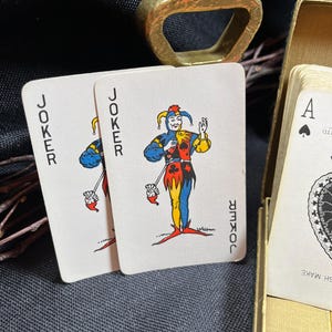 May include: Two playing cards with a joker illustration, featuring a jester in blue, yellow, and red attire. The word "JOKER" is printed vertically on each card. A gold-colored bottle opener and a deck of cards are also visible.