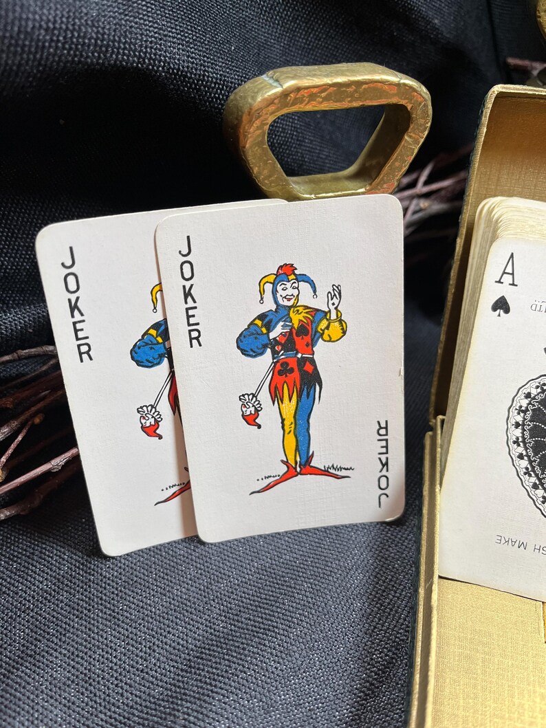 May include: Two playing cards with a joker illustration, featuring a jester in blue, yellow, and red attire. The word "JOKER" is printed vertically on each card. A gold-colored bottle opener and a deck of cards are also visible.