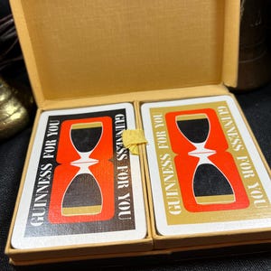 May include: A vintage deck of playing cards in a gold-colored box. The cards feature a red and black hourglass design with the text "GUINNESS FOR YOU" in white and gold. The box is open, revealing the cards.