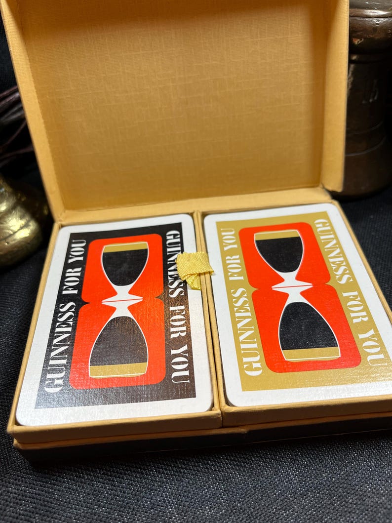 May include: A vintage deck of playing cards in a gold-colored box. The cards feature a red and black hourglass design with the text "GUINNESS FOR YOU" in white and gold. The box is open, revealing the cards.