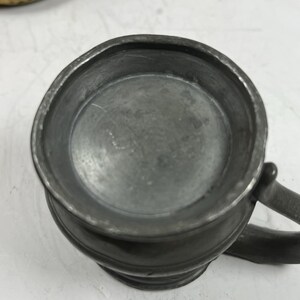 Puede incluir: Una peque&ntilde;a jarra de peltre antigua con asa. La jarra tiene un cuerpo redondeado y un borde ligeramente ensanchado. El peltre tiene un acabado gris mate. El asa es curva y est&aacute; unida al lateral de la jarra.