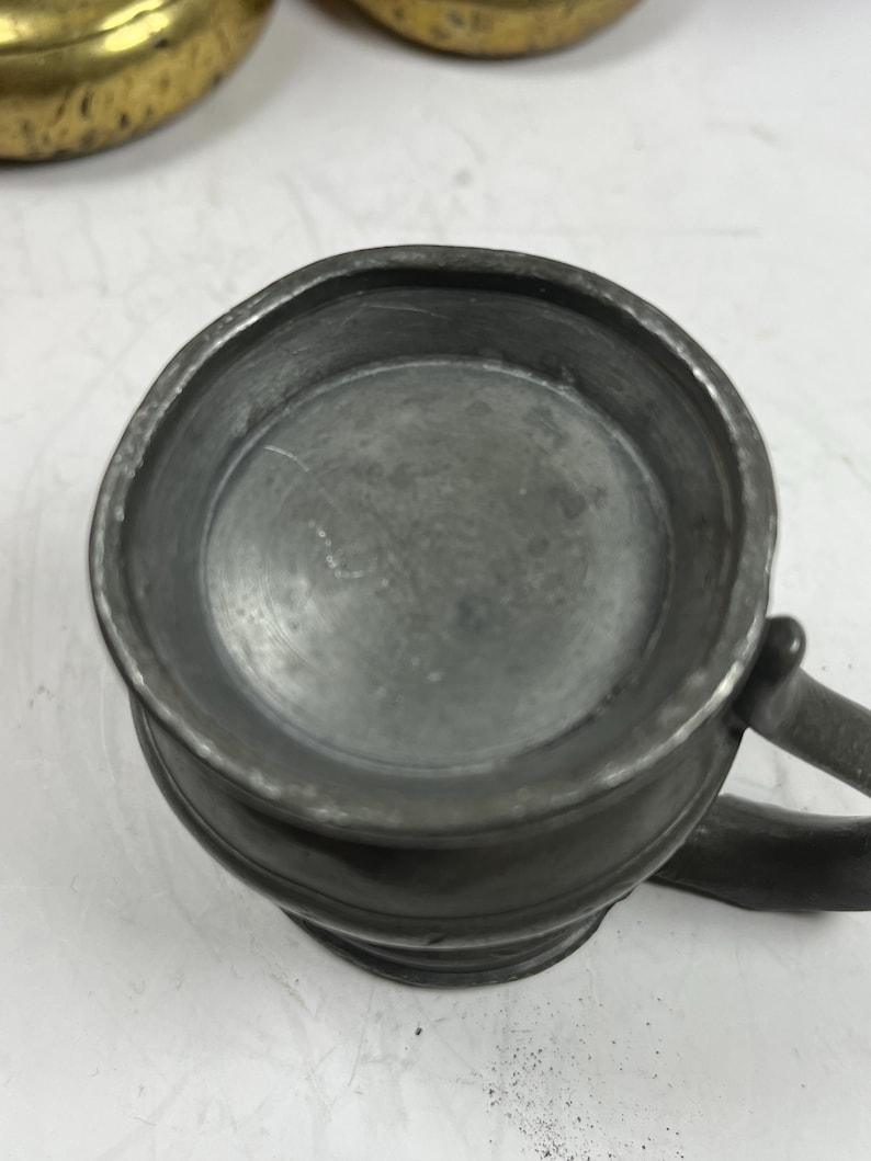 Puede incluir: Una peque&ntilde;a jarra de peltre antigua con asa. La jarra tiene un cuerpo redondeado y un borde ligeramente ensanchado. El peltre tiene un acabado gris mate. El asa es curva y est&aacute; unida al lateral de la jarra.