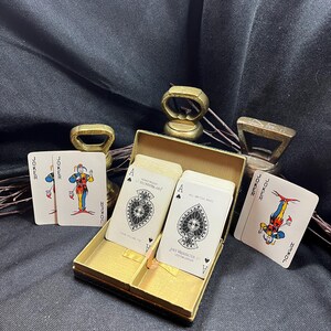 May include: A vintage playing card set in a gold-colored box, with two Jokers and two Ace of Spades cards visible. The cards feature detailed illustrations and text. The box is open, revealing the cards inside.