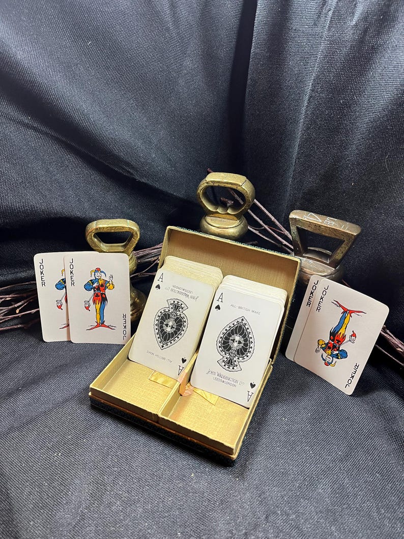 May include: A vintage playing card set in a gold-colored box, with two Jokers and two Ace of Spades cards visible. The cards feature detailed illustrations and text. The box is open, revealing the cards inside.
