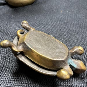 May include: A small, antique brass turtle-shaped trinket box. The turtle has a detailed shell and legs, with a hinged lid. The brass has a tarnished, aged appearance, with hints of green oxidation. The box is resting on a dark surface.