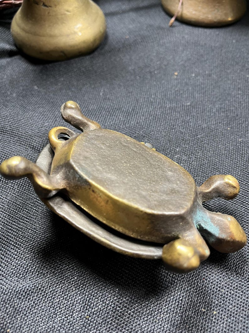 May include: A small, antique brass turtle-shaped trinket box. The turtle has a detailed shell and legs, with a hinged lid. The brass has a tarnished, aged appearance, with hints of green oxidation. The box is resting on a dark surface.