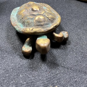 May include: A small, detailed bronze turtle figurine with a textured shell and four stubby legs. The turtle has a weathered appearance with hints of green patina, suggesting age. The figurine is a decorative item.