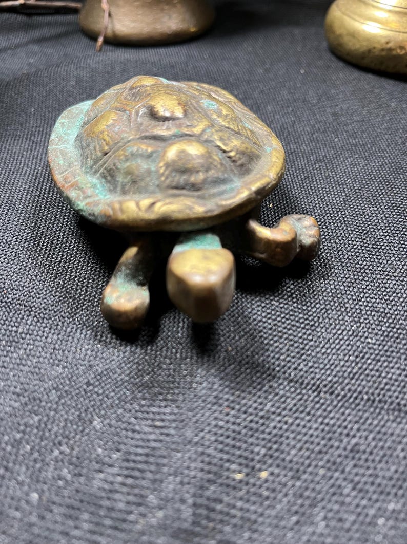 May include: A small, detailed bronze turtle figurine with a textured shell and four stubby legs. The turtle has a weathered appearance with hints of green patina, suggesting age. The figurine is a decorative item.