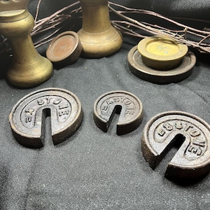 May include: Three cast iron slotted weights with the text "4 STONE" embossed on the surface, arranged in front of two brass bell-shaped weights and two circular brass weights. The iron weights are dark, textured, and feature a central slot.