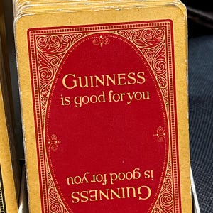 May include: A stack of vintage-style playing cards. Each card features a red oval with the text "GUINNESS is good for you" in gold lettering, framed by an ornate gold border. The cards have a gold edge.