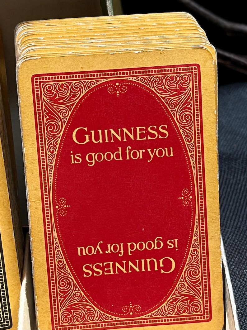 May include: A stack of vintage-style playing cards. Each card features a red oval with the text "GUINNESS is good for you" in gold lettering, framed by an ornate gold border. The cards have a gold edge.
