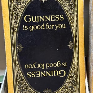 May include: A rectangular card with a gold border and a black oval in the center. The text "GUINNESS is good for you" is printed in gold. The same text is also printed upside down. The card has an ornate design.