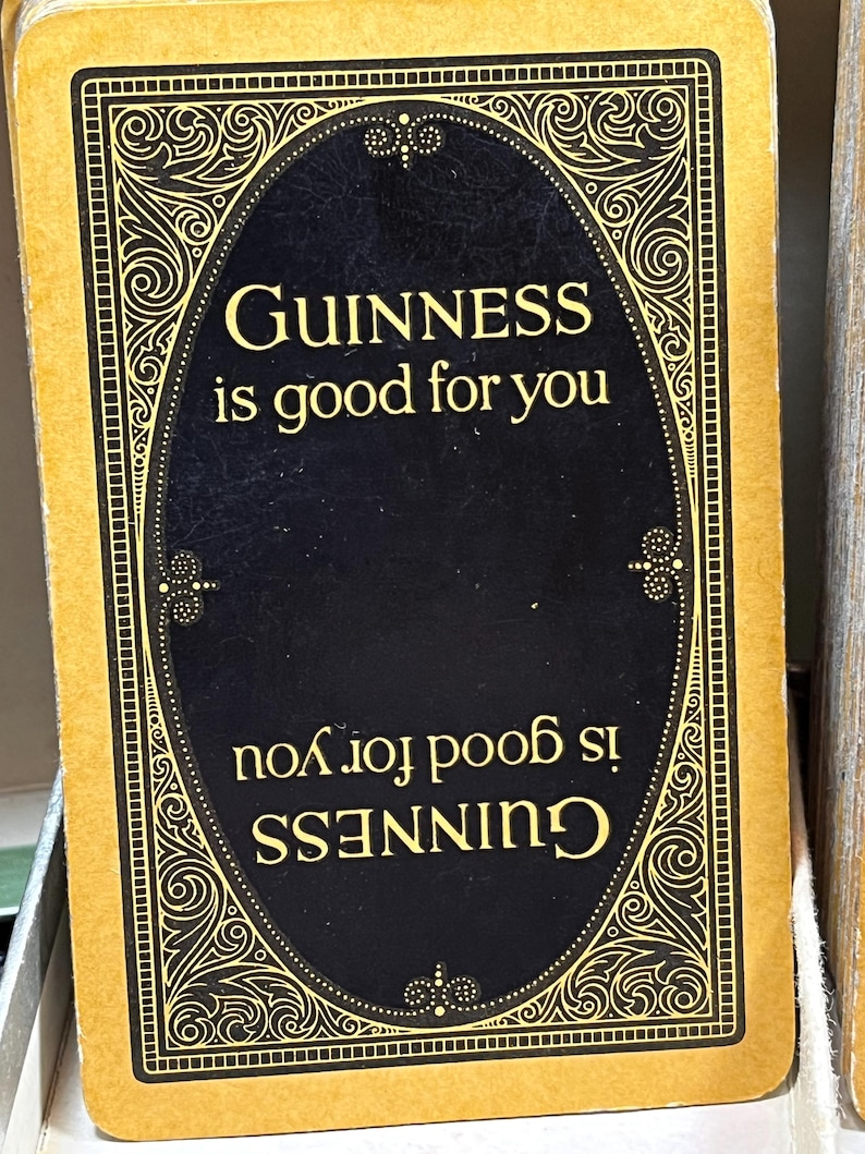 May include: A rectangular card with a gold border and a black oval in the center. The text "GUINNESS is good for you" is printed in gold. The same text is also printed upside down. The card has an ornate design.
